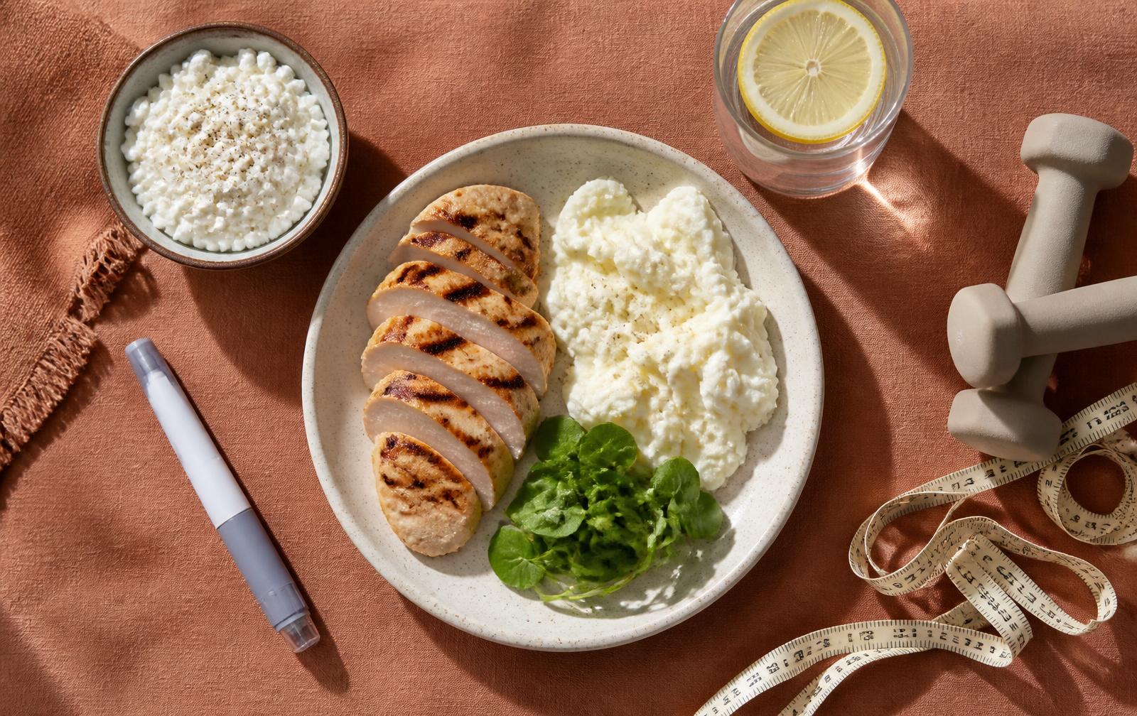 Overhead flat lay on terracotta linen of grilled chicken slices and egg whites on a plate, a bowl of cottage cheese, a glass of lemon water, light dumbbells, a tape measure, and an unbranded injection pen.